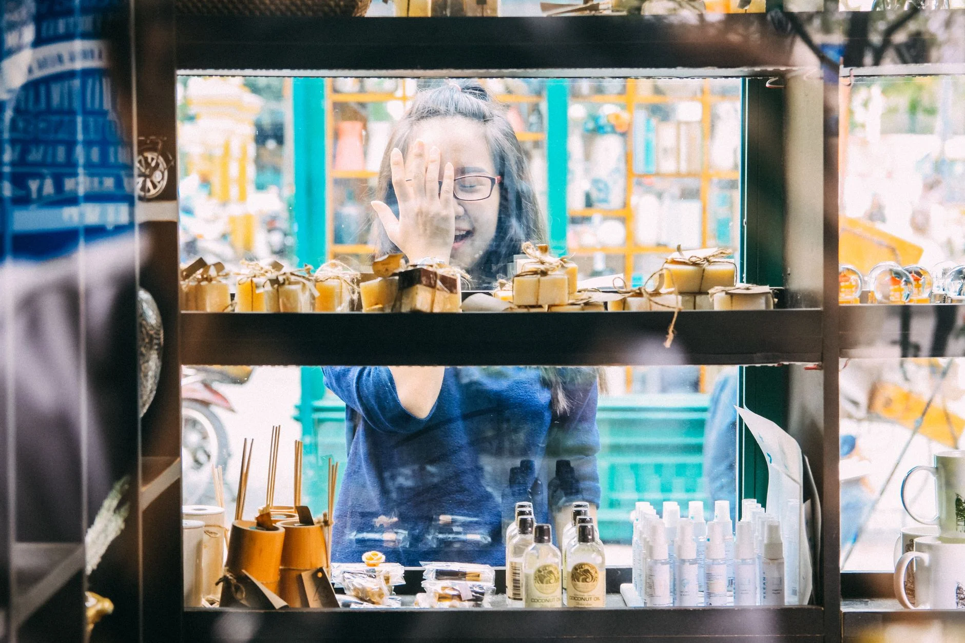 A cheerful woman peering through a glass display filled with boutique soaps and bottles.