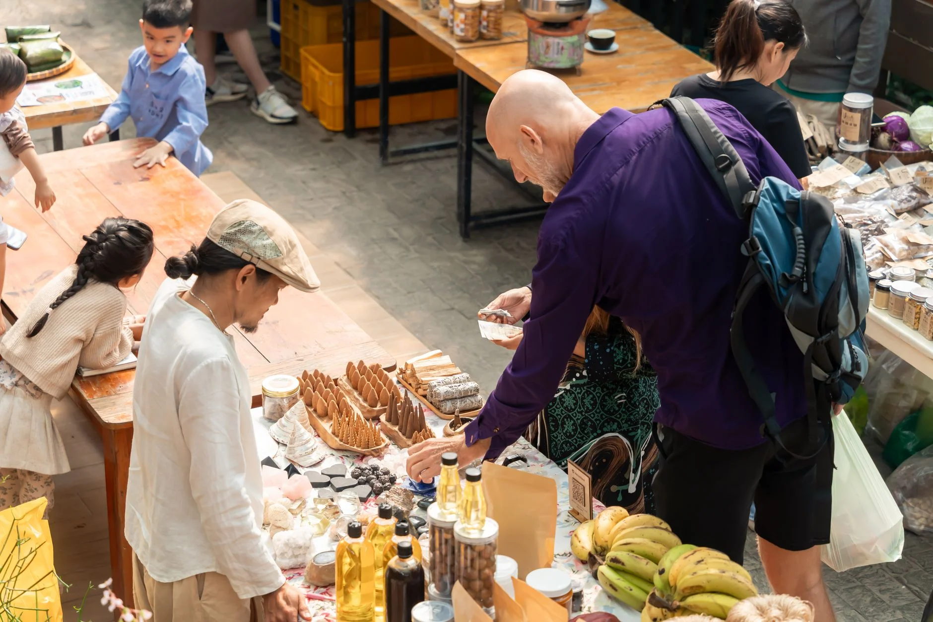 A vibrant outdoor market with diverse people browsing and buying assorted products from vendors.